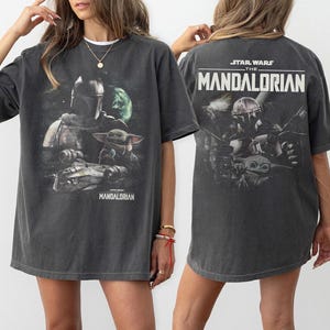 May include: Dark gray t-shirt featuring a graphic from The Mandalorian. The front design includes the Mandalorian, Grogu, and a spaceship. The back of the shirt displays the Star Wars logo and the words "The Mandalorian".