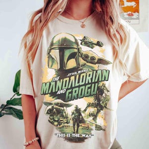 May include: Cream-colored t-shirt featuring a graphic from the Star Wars series, "The Mandalorian and Grogu." The design includes images of the Mandalorian, Grogu, and a spaceship, with the phrase "THIS IS THE WAY" at the bottom.