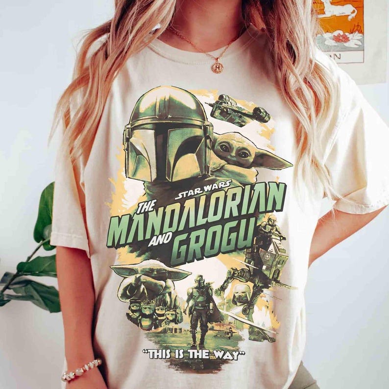 May include: Cream-colored t-shirt featuring a graphic from the Star Wars series, "The Mandalorian and Grogu." The design includes images of the Mandalorian, Grogu, and a spaceship, with the phrase "THIS IS THE WAY" at the bottom.