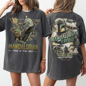 May include: Dark gray t-shirt featuring a graphic of the Mandalorian and Grogu. The front of the shirt displays the Mandalorian and Grogu with the text "MANDALORIAN - THIS IS THE WAY." The back of the shirt has the text "STAR WARS THE MANDALORIAN AND GROGU - THIS IS THE WAY."