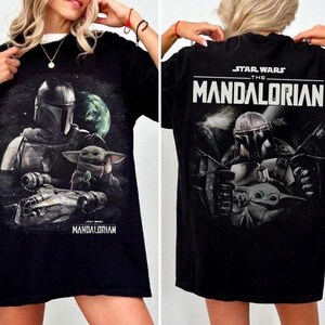 May include: Black t-shirt featuring a graphic of The Mandalorian and Grogu from Star Wars. The front of the shirt shows the characters and a spaceship, while the back displays the show's title. The shirt has a white collar.