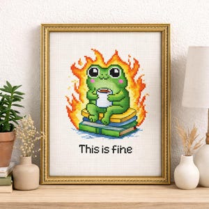 May include: Cross-stitch art in a gold frame featuring a green frog with a cup, sitting on books, surrounded by flames. The text "This is fine" is below the frog. The artwork is displayed on a wooden surface.