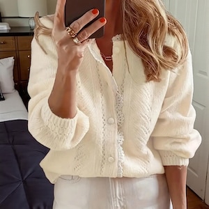 Könnte beinhalten: Cremefarbene Strickjacke mit zarter Spitzenborte und Knopfleiste. Der langärmlige Pullover hat ein strukturiertes Strickmuster und wird mit weißen Jeans kombiniert. Das Outfit ist ein lässiger, alltäglicher Stil.
