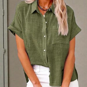 May include: Olive green short-sleeved button-down shirt with a chest pocket. The shirt has a collared neckline and is paired with white pants. The fabric appears to have a textured weave.