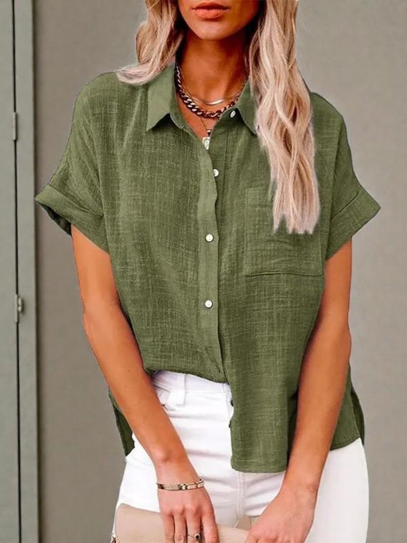 May include: Olive green short-sleeved button-down shirt with a chest pocket. The shirt has a collared neckline and is paired with white pants. The fabric appears to have a textured weave.