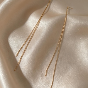 May include: A pair of delicate gold-colored earrings. Each earring features a long, slender, straight bar that curves slightly at the end. The earrings are displayed on a satin fabric.
