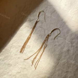 Peut inclure: Une paire de boucles d'oreilles pendantes dorées. Chaque boucle d'oreille est dotée d'un crochet incurvé et de plusieurs brins de chaînes délicates qui tombent en cascade. Les boucles d'oreilles sont présentées sur une surface texturée blanc cassé.