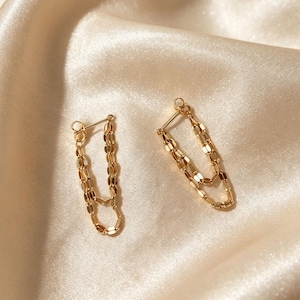 May include: A pair of gold chain earrings. Each earring has a post and two delicate chains made of small, rectangular links. The earrings are set against a soft, cream-coloured satin background, creating a luxurious aesthetic.