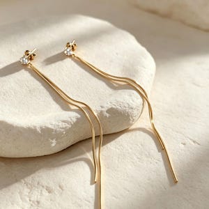 May include: A pair of gold-toned drop earrings featuring a small, clear, round-cut gemstone stud at the top. Each earring consists of two delicate, thin, wavy gold chains that hang downward from the stud, creating a fluid and elongated appearance.