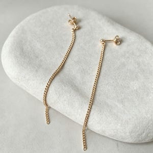 Puede incluir: Un par de pendientes de cadena dorada con un pequeño poste de perno con un detalle de bola y una delicada cadena de eslabones que cuelga verticalmente, creando un diseño lineal simple y elegante para las orejas.