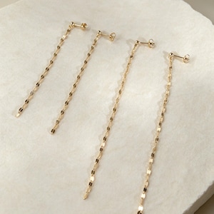 Peut inclure: Quatre boucles d'oreilles pendantes dorées, chacune composée d'une chaîne délicate faite de petits maillons circulaires plats et réfléchissants. Les boucles d'oreilles sont des clous avec une petite bille au sommet, laissant la chaîne pendre.