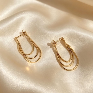 May include: A pair of gold-coloured earrings, each with a layered teardrop shape. The earrings have three delicate, curved strands. They are presented on a cream-coloured, satin-like fabric.