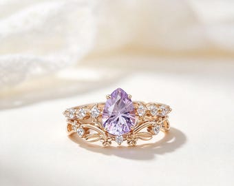 Può includere: Un anello in oro rosa con una pietra preziosa centrale color viola chiaro a forma di goccia. La fascia è decorata con piccole pietre trasparenti e motivi metallici a forma di foglie e fiori che incorniciano la pietra principale.