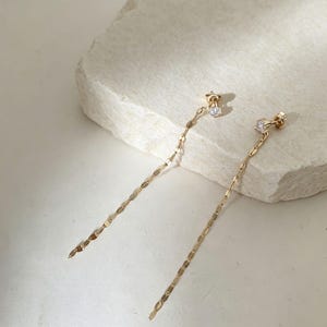 May include: A pair of gold drop earrings featuring a small, clear, round-cut gemstone stud at the top. Each earring has a long, delicate chain composed of small, flat, oval-shaped gold links that hang vertically from the gemstone setting.