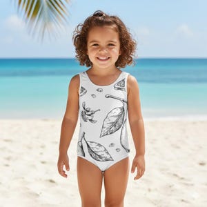Op de afbeelding: Een witte badpak met een zwart-wit botanische illustratie van citroenen, bladeren en een insect. Het mouwloze badpak heeft een ronde hals. Het kind staat op een zandstrand met de zee op de achtergrond.