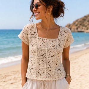 May include: Cream-colored crochet top with a square neckline and short sleeves. The top features a geometric pattern and is paired with light-colored shorts. The outfit is worn on a beach setting, suggesting a summer or vacation theme.