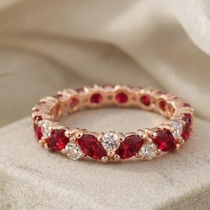 May include: A rose gold eternity band featuring alternating marquise-cut red gemstones and round-cut clear white stones. The ring is positioned on a textured block, with the text Jewels Aroh Jewels Aroh Jewels visible at the top of the frame.