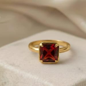 Pode incluir: Um anel de ouro com uma pedra preciosa vermelha escura quadrada. O anel está sobre uma superfície branca texturizada, com um tecido branco e macio no fundo. É uma joia.