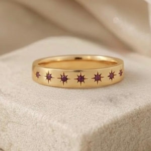May include: A gold-colored ring with a series of small, dark red star-shaped designs. The ring is set on a small, square, off-white block. The background features soft, white fabric and small white flowers. The text "Aroh Jewels Aroh Jewels Aroh" is at the top.