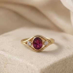 May include: A gold ring featuring an oval, deep pink gemstone, flanked by two smaller, round, clear gemstones. The ring is set on a neutral-colored, textured surface, with a soft, white fabric in the background. The text "Aroh Jewels" is visible.