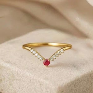 May include: A gold-colored ring with a V-shaped design. The ring features small, clear stones along the V's arms and a single, round, red stone at the point. The ring is set on a square, light-colored surface.