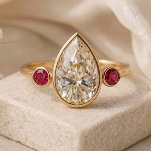 Puede incluir: Un anillo de oro con una piedra preciosa clara en forma de pera y dos piedras rojas redondas más pequeñas. El anillo está sobre una superficie texturizada de color neutro. El anillo es una joya.