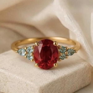 May include: A gold ring with a large, oval-cut, deep red gemstone as the focal point. It's flanked by smaller, light blue gemstones. The ring is displayed on a white surface, against a soft, neutral-toned backdrop.