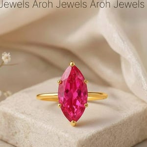 May include: A gold ring featuring a marquise-cut, vibrant pink gemstone. The ring is set on a small, textured, rectangular block. The background is a soft, neutral color, with delicate white flowers and the text "Aroh Jewels" visible.