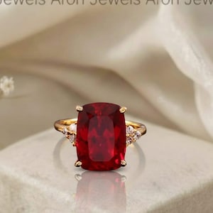 May include: A gold ring with a large, rectangular, deep red gemstone as the centerpiece. The ring has smaller, clear gemstones on either side of the main stone. The ring is set on a white, textured surface.
