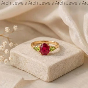 Può includere: Un anello d'oro con una grande pietra centrale di rubino ovale, affiancata da due gemme verdi a forma di pera. L'anello è posto su una superficie chiara e strutturata. Il marchio "Aroh Jewels" è ripetuto in alto.