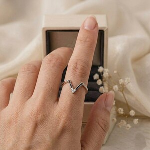 Op de afbeelding: Een zilveren V-vormige ring met zwarte en witte stenen. De ring zit om een vinger, met een sieradendoosje op de achtergrond. De ring heeft een modern, minimalistisch ontwerp.