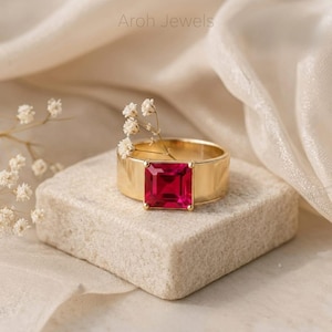 May include: A gold ring with a wide band and a square, vibrant red gemstone. The ring is displayed on a small, textured, light-colored block, with delicate white flowers and soft, cream-colored fabric in the background. The text "Aroh Jewels" is visible.