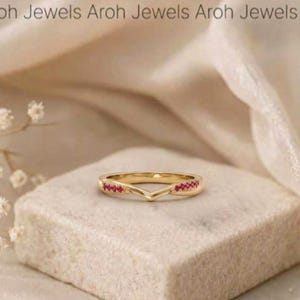 May include: A gold V-shaped ring featuring a series of small, vibrant red stones set along the band. The text Aroh Jewels is visible at the top of the frame. The ring has a polished metallic finish and a pointed chevron design.