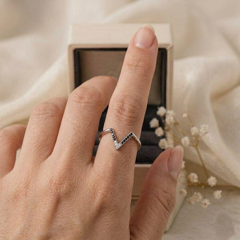 Op de afbeelding: Zilveren V-vormige ring met zwarte en witte stenen. De ring zit om een vinger, met een sieradendoosje op de achtergrond. De ring heeft een modern, minimalistisch ontwerp.