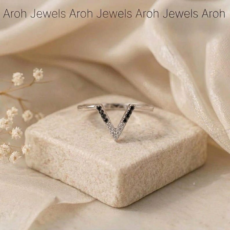 Op de afbeelding: Een zilveren ring met een V-vormig ontwerp. De V is gevuld met kleine zwarte en witte stenen. De ring wordt gepresenteerd op een kleine, lichtgekleurde stenen blok, met de tekst "Aroh Jewels" bovenaan.
