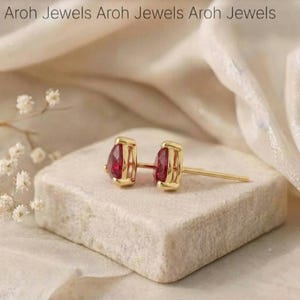 Puede incluir: Un par de pendientes de oro con piedras preciosas rojas en forma de pera en un engaste de garras. El texto Aroh Jewels Aroh Jewels Aroh Jewels es visible en la parte superior. Los pendientes están sobre un bloque de piedra de color claro.