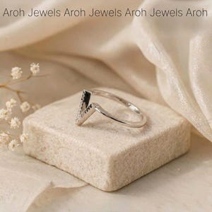 Op de afbeelding: Een zilveren ring met een V-vormig ontwerp, bezet met kleine, sprankelende stenen. De ring ligt op een kleine, vierkante, beige steen. De achtergrond is een zachte, cr&egrave;mekleurige stof. De tekst "Aroh Jewels" staat bovenaan.