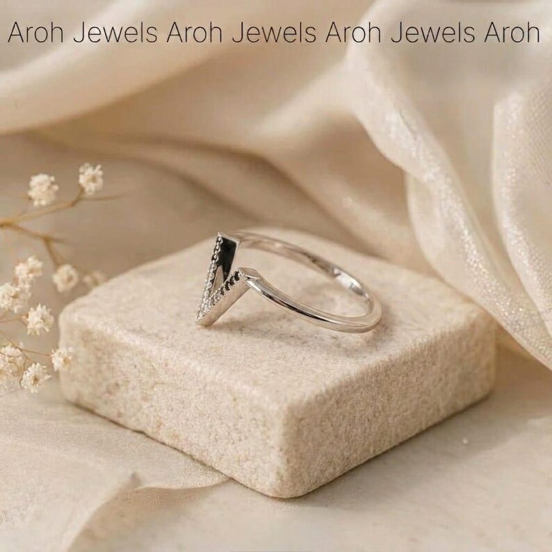 Op de afbeelding: Een zilveren ring met een V-vormig ontwerp, bezet met kleine, sprankelende stenen. De ring ligt op een kleine, vierkante, beige steen. De achtergrond is een zachte, cr&egrave;mekleurige stof. De tekst "Aroh Jewels" staat bovenaan.
