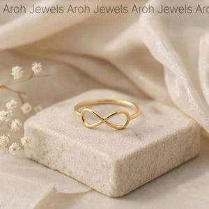 May include: A gold-toned ring featuring a central infinity symbol design. The ring is positioned on a textured, light-colored stone block. The text Aroh Jewels is repeated across the top of the image in a simple, dark, sans-serif font.