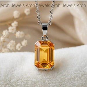 May include: A silver pendant necklace featuring a rectangular, faceted, golden-yellow gemstone. The pendant is suspended from a delicate chain. The jewelry is set against a soft, neutral background.