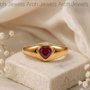 May include: A gold ring featuring a central heart-shaped red gemstone. The band is smooth and polished, tapering slightly towards the setting. The text Aroh Jewels is visible at the top and bottom of the image, identifying the brand name clearly.