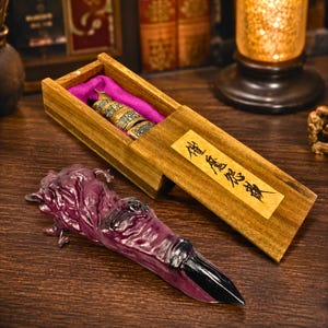 May include: A dark purple, textured, claw-shaped object with a black pointed tip, positioned next to a rectangular wooden box. The box features a sliding lid with a paper label containing black Chinese characters and a purple fabric-lined interior.