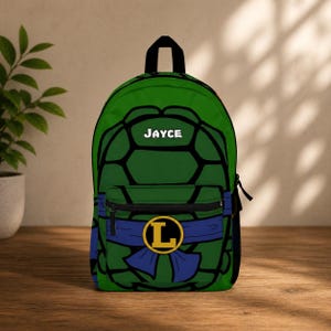 Könnte beinhalten: Grüner Rucksack mit schwarzem Umrissdesign, mit dem Namen "JAYCE" in Weiß. Der Rucksack hat einen blauen Schärpe mit einem gelben "L"-Emblem und eine Vordertasche. Die Tasche steht auf einer Holzoberfläche.