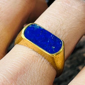 Könnte beinhalten: Ein goldfarbener Ring mit einem rechteckigen, tiefblauen Stein in der Mitte. Der Ring hat ein schlichtes, klassisches Design, und der Stein hat ein leicht strukturiertes Aussehen.