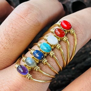 Può includere: Una collezione di anelli color oro, ognuno con una gemma ovale di colore diverso. Le gemme sono di vari colori, tra cui rosso, bianco, turchese, blu e viola. Gli anelli sono esposti su un dito.