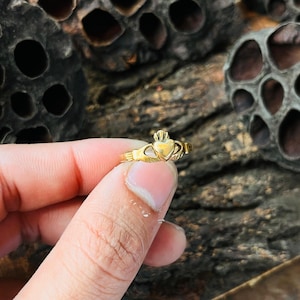 May include: A gold Claddagh ring with a heart, crown, and hands design. The ring is small and detailed. The ring is held up against a dark, textured background.