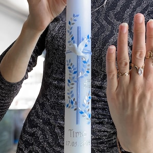 Könnte beinhalten: Eine weiße Kerze mit blauen und silbernen Blatt- und Kreuzmotiven, einer weißen Taube und dem Namen "Timo" mit dem Datum "17.05.2026". Die Kerze wird von einer Person gehalten.