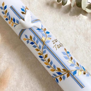 Könnte beinhalten: Eine weiße zylindrische Kerze, verziert mit einer dreidimensionalen weißen Taube, blau-goldenen Rankenmustern und einem blauen Kreuz. Der Schriftzug "zur Taufe" ist in goldener Schrift auf der Seite der glatten Kerze angebracht.