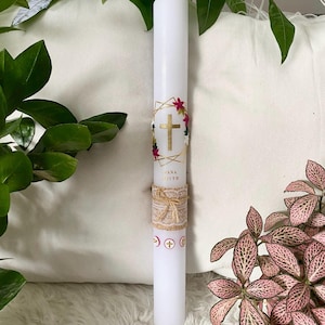 Könnte beinhalten: Eine weiße Säulenkerze mit einem goldenen Kreuzdesign, floralen Akzenten und dem Namen "Jana Faith". Die Kerze ist mit einem Jutegürtel und Kreuzsymbolen verziert. Grüne Pflanzen und rosa Blüten umgeben die Kerze.