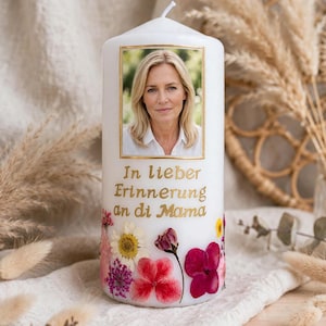 May include: A white cylindrical pillar candle featuring a rectangular photograph of a blonde person framed in gold. Below the image, the gold text reads In lieber Erinnerung an di Mama. The base is decorated with various colourful pressed flowers.